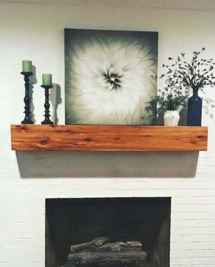 Red cedar mantle with decor, featuring candlesticks and artwork, installed above a white brick fireplace.