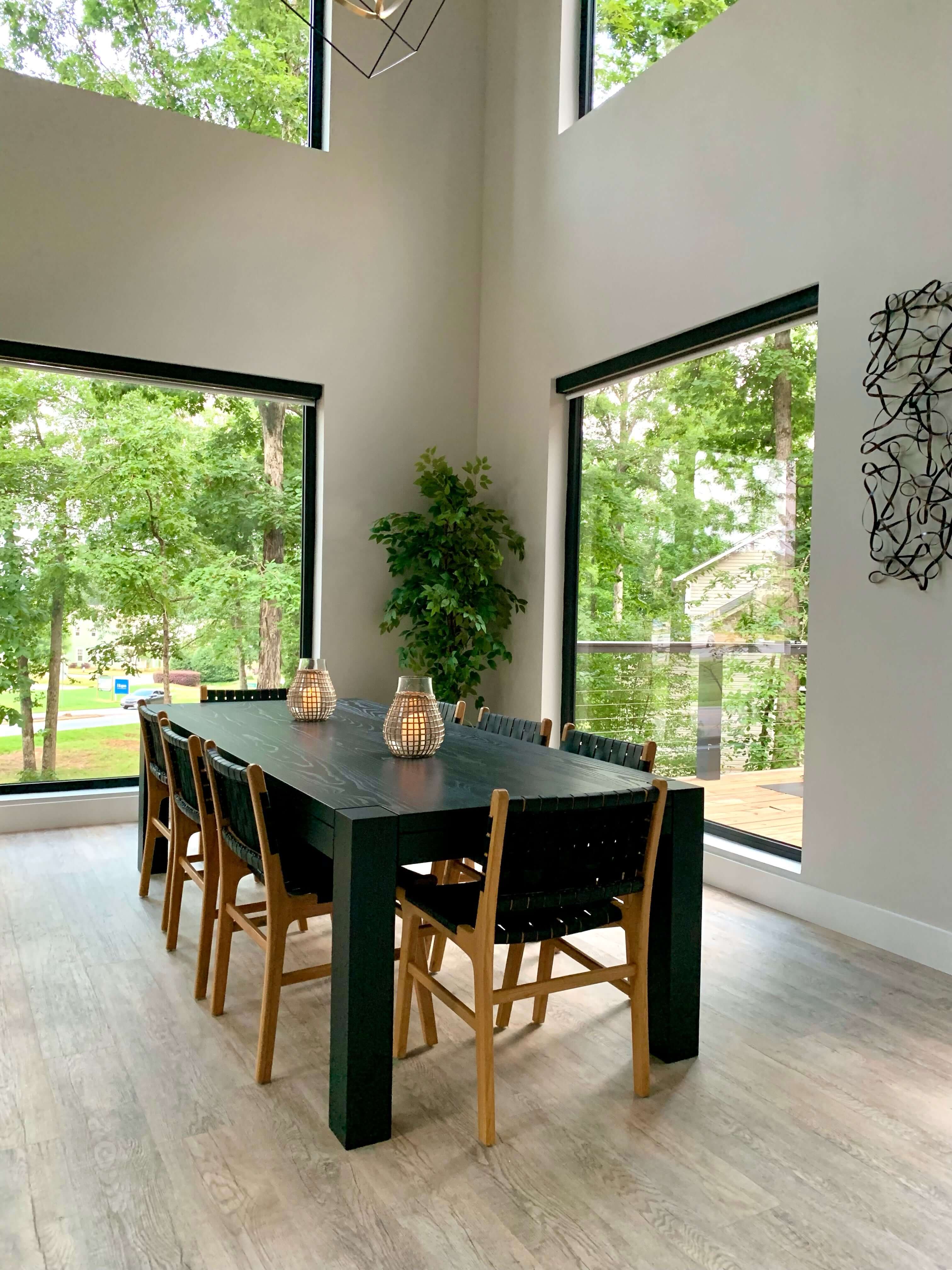 Modern dining room featuring a Royal Flush Dining Table with sleek black finish, natural light, and large windows offering a forest view.