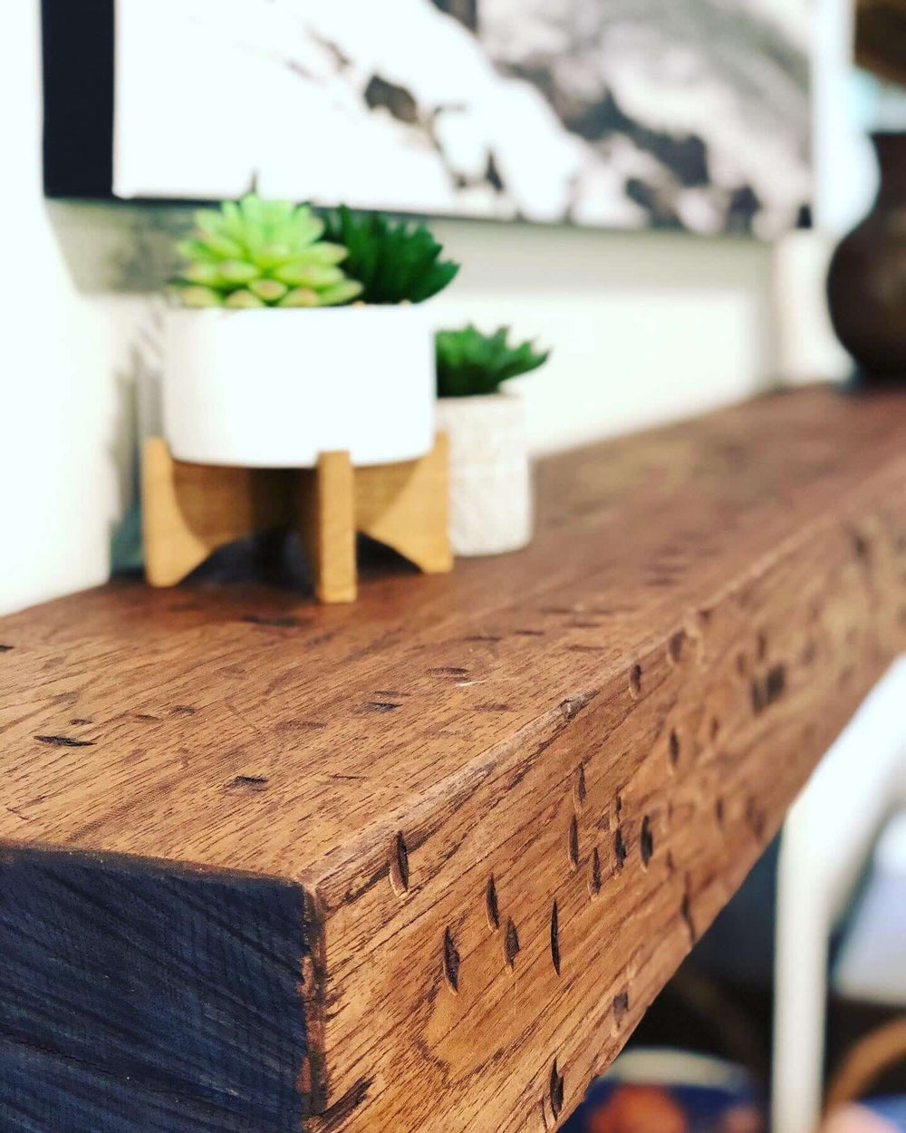 Rustic solid wood hand-hewn mantel with dark walnut stain, featuring decorative plants, enhancing a cozy living space.