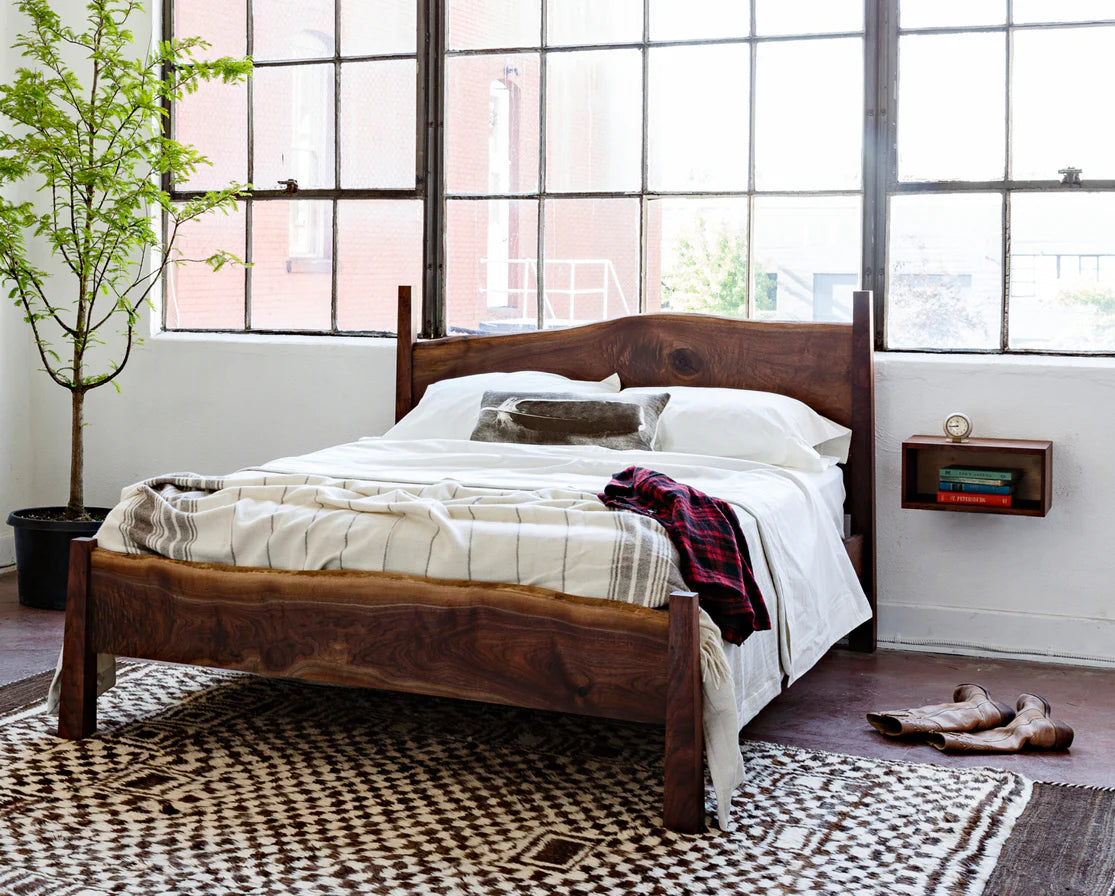 Wooden bed in a room with large windows and a plant.